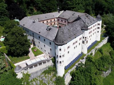 An aerial view of a large, white-walled castle complex with grey slate roofs, situated on a rocky outcrop surrounded by dense green forest, featuring prominent corner towers and solar panels installed on the lower walls.