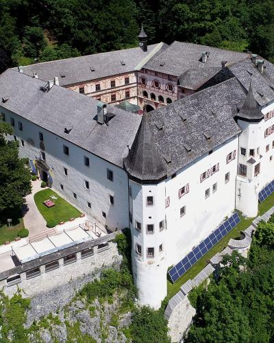 An aerial view of a large, white-walled castle complex with grey slate roofs, situated on a rocky outcrop surrounded by dense green forest, featuring prominent corner towers and solar panels installed on the lower walls.