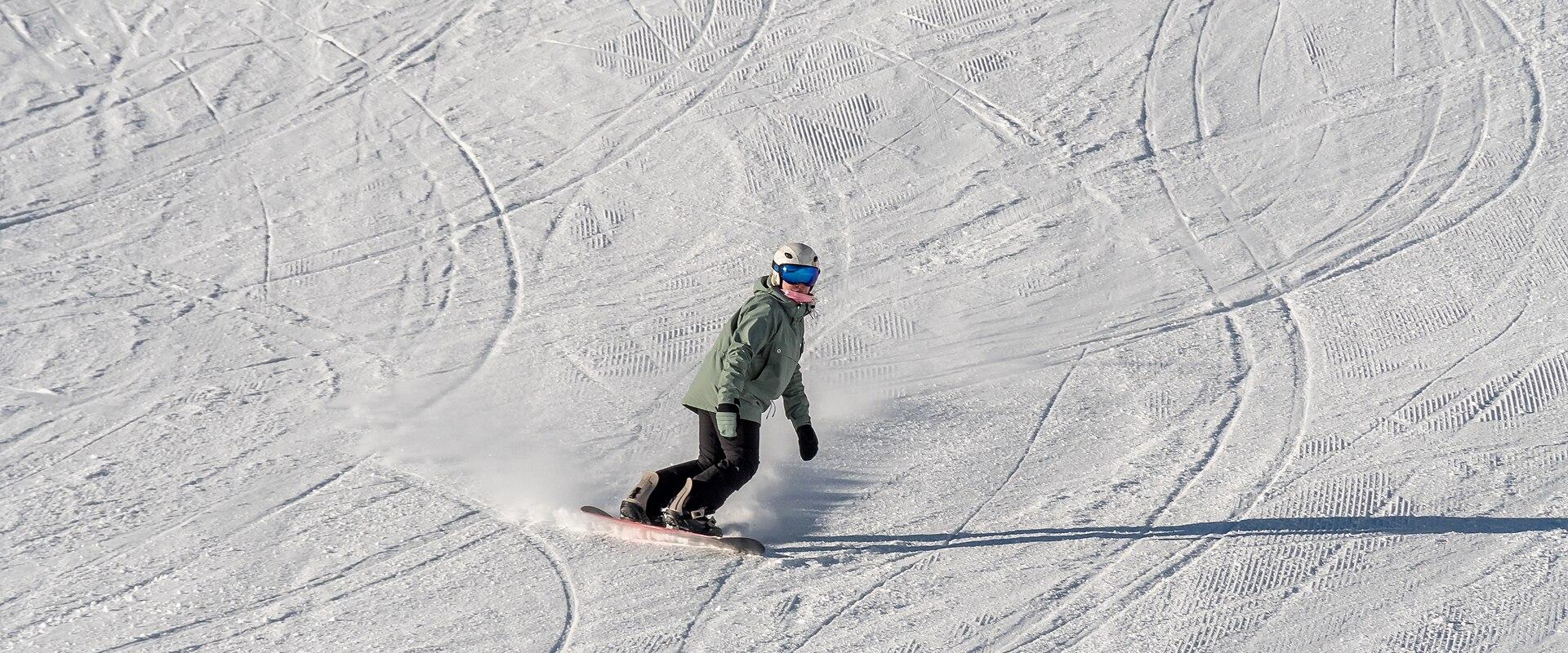 A snowboarder in a green jacket and helmet descends a groomed, snow-covered slope marked with curved tracks and parallel grooming lines, under bright sunlight.