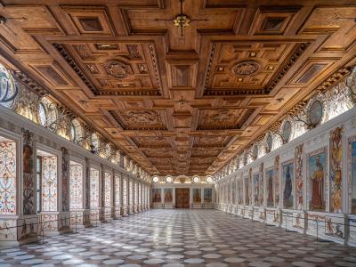 A long, ornate interior hall featuring a highly detailed wooden coffered ceiling, white walls decorated with numerous painted portraits and grotesque motifs, and a geometric tiled floor.