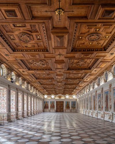 A long, ornate interior hall featuring a highly detailed wooden coffered ceiling, white walls decorated with numerous painted portraits and grotesque motifs, and a geometric tiled floor.