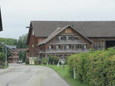 A traditional alpine farmhouse with a large wooden barn section and a residential gable front, situated along a paved village road with a house number sign reading "10" and a car in the distance.
