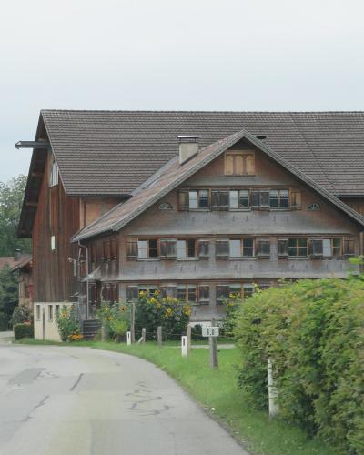 A traditional alpine farmhouse with a large wooden barn section and a residential gable front, situated along a paved village road with a house number sign reading "10" and a car in the distance.