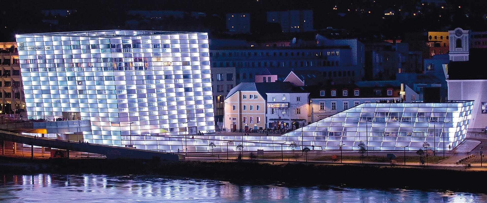 The image shows the Ars Electronica Center in Linz, Austria, a modern glass-and-steel building illuminated at night along the Danube River, with its distinctive grid-like facade and adjacent lower structure visible.