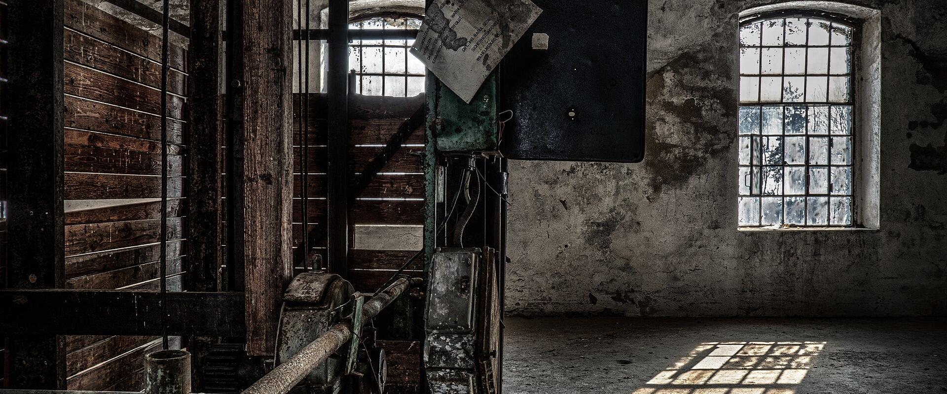 An interior shot of a derelict industrial space featuring heavy machinery, peeling walls, arched windows, and a hanging sign that reads "Gang 4".