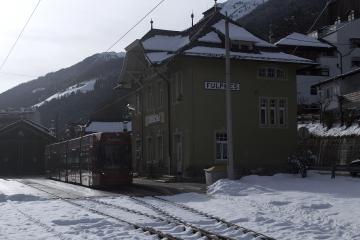A red tram or light rail vehicle on tracks in a snowy alpine setting, with a green station building labeled "FULPMES" and mountainous terrain in the background.