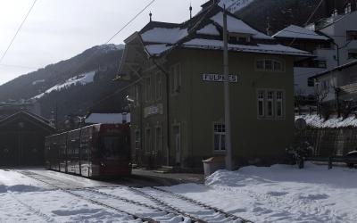 A red tram or light rail vehicle on tracks in a snowy alpine setting, with a green station building labeled "FULPMES" and mountainous terrain in the background.