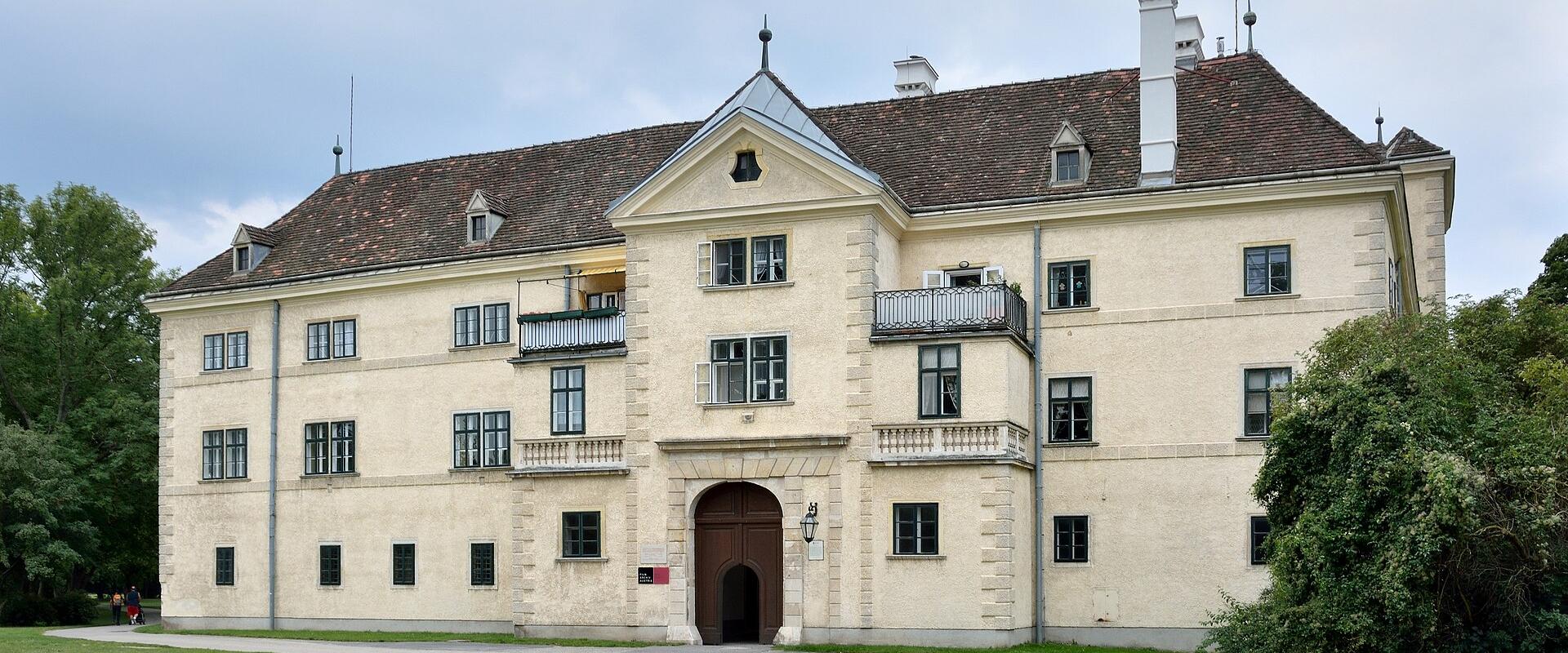 A large, pale-yellow historic building with a central arched entrance, balconies, and a tiled roof, set in a grassy park-like setting with trees; signage near the entrance appears to read "Filmarchiv Austria" or similar institutional text.