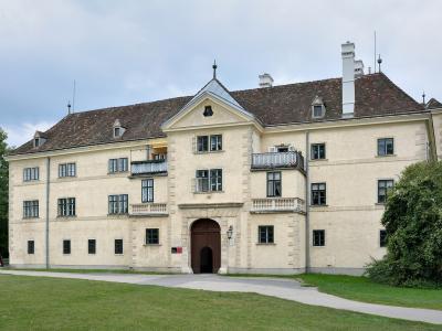 A large, pale-yellow historic building with a central arched entrance, balconies, and a tiled roof, set in a grassy park-like setting with trees; signage near the entrance appears to read "Filmarchiv Austria" or similar institutional text.