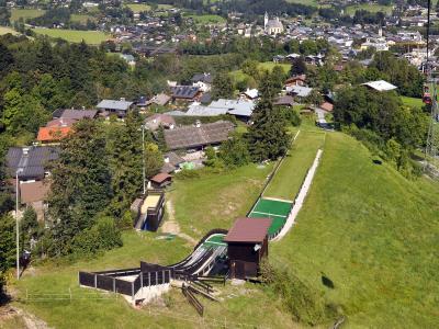An aerial view of a ski jump facility on a grassy hillside with a town and church in the background, featuring a long green landing slope and a cable car tower to the right.