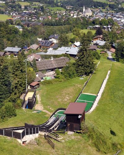 An aerial view of a ski jump facility on a grassy hillside with a town and church in the background, featuring a long green landing slope and a cable car tower to the right.