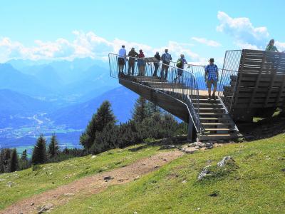 A wooden viewing platform with metal railings and stairs situated on a grassy mountain slope, with several people standing on it and a panoramic view of distant blue mountains and a valley below.