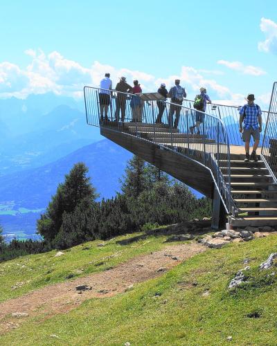 A wooden viewing platform with metal railings and stairs situated on a grassy mountain slope, with several people standing on it and a panoramic view of distant blue mountains and a valley below.