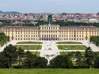 A wide-angle view of a large, yellow Baroque palace with formal gardens and a cityscape in the background.
