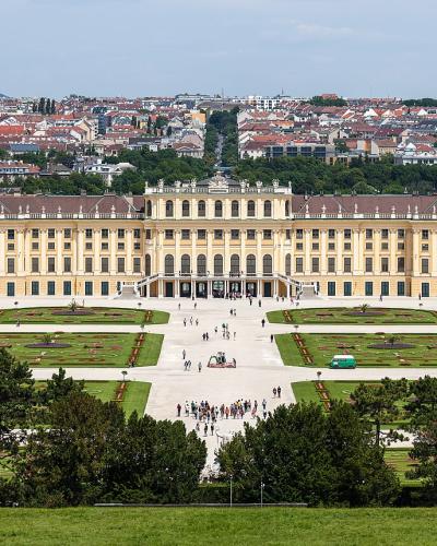 A wide-angle view of a large, yellow Baroque palace with formal gardens and a cityscape in the background.
