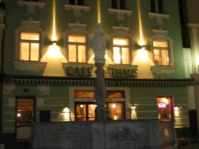 A night photograph of a stone fountain with a statue in the foreground, situated in front of a green building with a sign reading "CAFE RATHAUS".