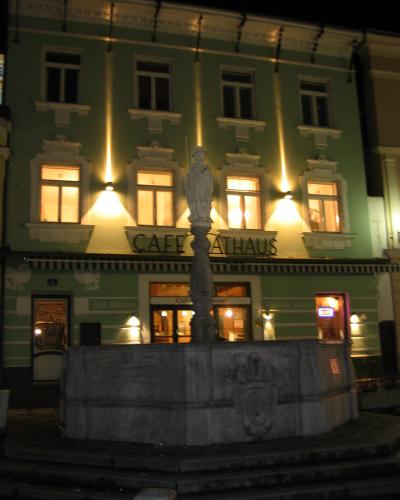 A night photograph of a stone fountain with a statue in the foreground, situated in front of a green building with a sign reading "CAFE RATHAUS".