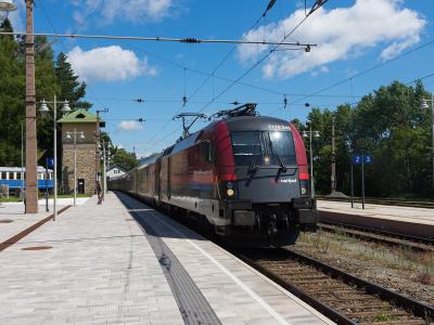 A red and grey ÖBB Railjet train stopped at an outdoor railway station platform with overhead catenary lines, a stone signal box, and blue platform signage.