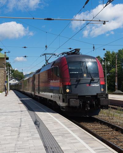 A red and grey ÖBB Railjet train stopped at an outdoor railway station platform with overhead catenary lines, a stone signal box, and blue platform signage.