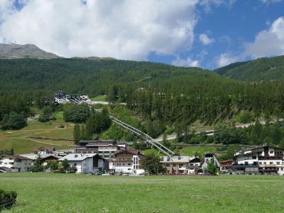 A wide landscape shot of the village of Sölden in the Ötztal Alps, showing traditional alpine buildings in the foreground and a cable car line ascending the forested mountain slope towards the Rettenbach glacier area.