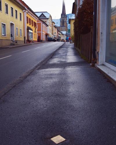 A wet street scene in a European town featuring a brass cobblestone memorial plaque (Stolperstein) in the foreground, colorful buildings along the road, and a church spire in the distance.