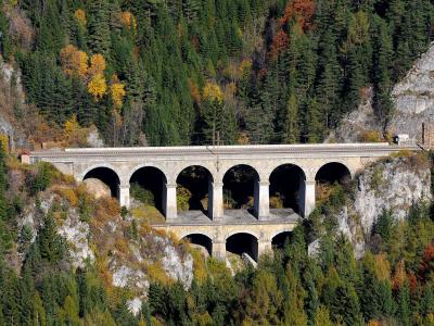 A stone railway viaduct with two tiers of arches set into a steep, forested mountainside with autumn foliage.