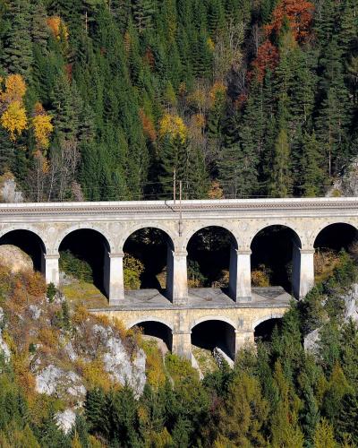 A stone railway viaduct with two tiers of arches set into a steep, forested mountainside with autumn foliage.