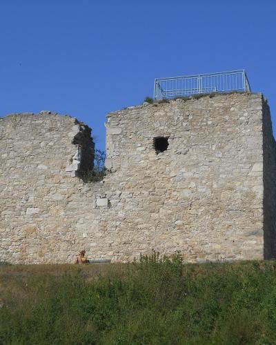 The image shows a stone ruin on a grassy hill, featuring a large, partially collapsed wall with a modern metal safety railing installed on the top edge, set against a clear blue sky.