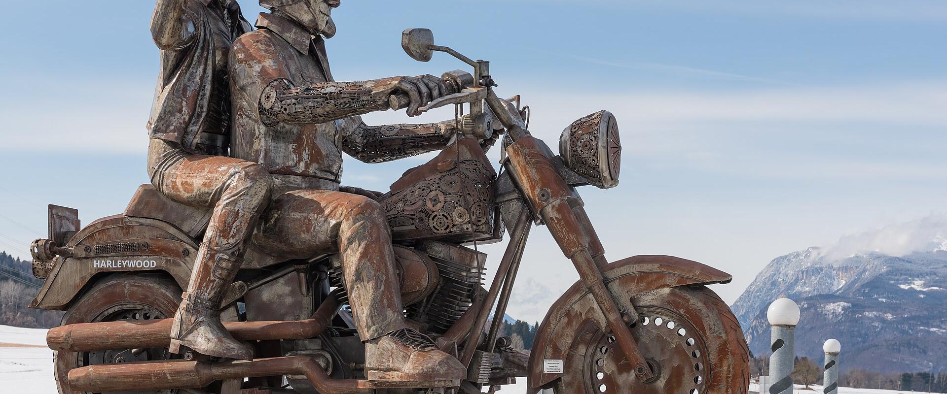 A large, rusted metal sculpture of two figures riding a motorcycle, with "HARLEYWOOD" visible on the rear fender, situated at a roadside roundabout with snow-covered ground and mountains in the background.