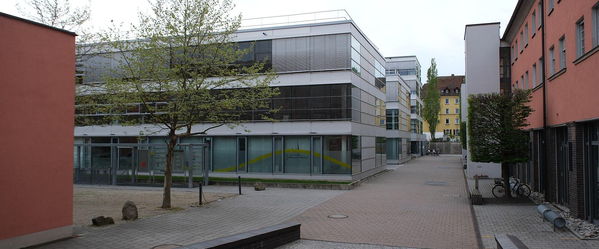 The image shows a modern multi-story building with glass and metal cladding, situated in a paved courtyard with trees, benches, and adjacent pinkish-red buildings; signage on the glass facade appears to read "Posthof" with a yellow arc design.