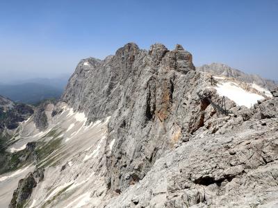 A high-altitude view of a rugged, rocky mountain ridge with patches of snow and a metal staircase or ladder structure built into the rock face on the right side.