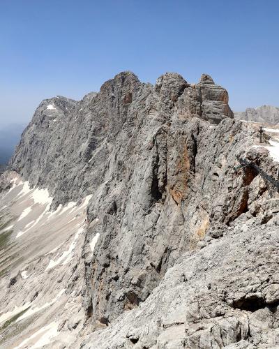A high-altitude view of a rugged, rocky mountain ridge with patches of snow and a metal staircase or ladder structure built into the rock face on the right side.