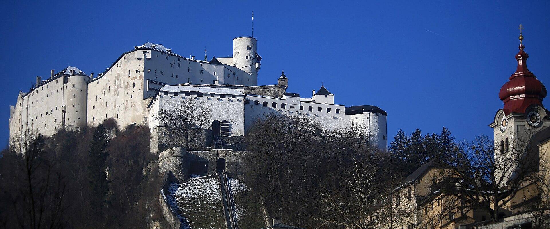 A wide-angle photograph of the Hohensalzburg Fortress on a hill, with a funicular railway track (Reißzug) ascending the slope below it, and the red-domed tower of Stift Nonnberg visible in the foreground right.