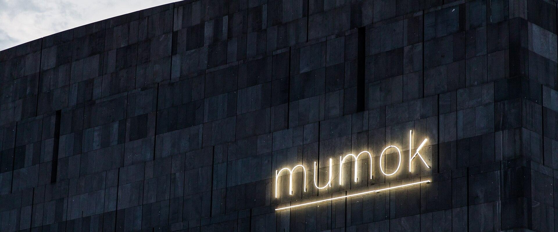 The image shows the illuminated "mumok" sign on the dark, tiled facade of a modern building under an overcast sky.