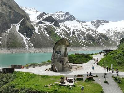 A large, curved concrete monument stands on a paved plaza beside a turquoise alpine reservoir, with snow-dusted mountains in the background and visitors walking nearby.