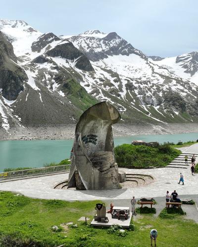 A large, curved concrete monument stands on a paved plaza beside a turquoise alpine reservoir, with snow-dusted mountains in the background and visitors walking nearby.