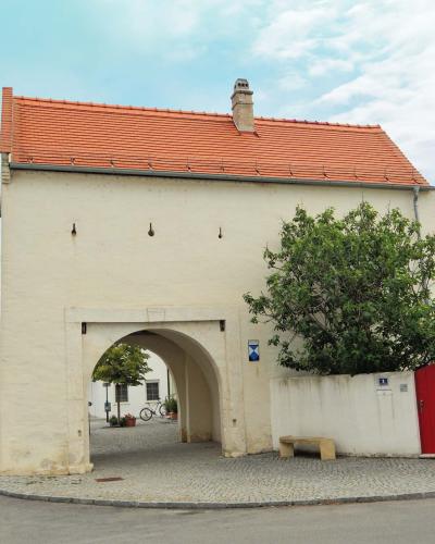 A white, two-story building with a red tiled roof, featuring a large arched passageway on the ground floor, a red gate to the right, and a blue and white sign on the wall.