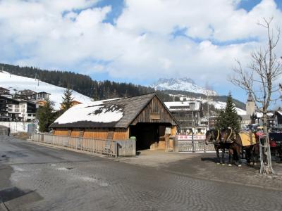 A wooden covered bridge structure with a snow-dusted roof, situated next to a cobblestone road with horses and carriages nearby, set against a backdrop of snowy mountains and alpine buildings.