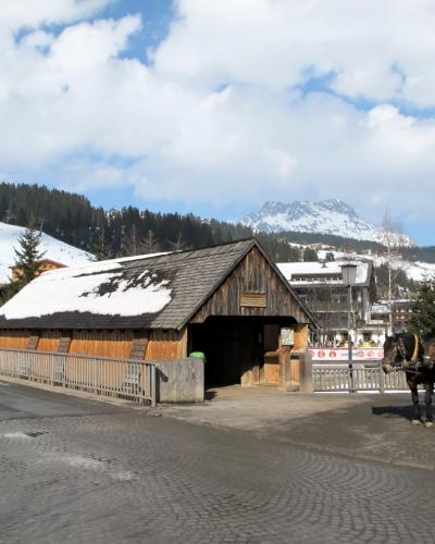 A wooden covered bridge structure with a snow-dusted roof, situated next to a cobblestone road with horses and carriages nearby, set against a backdrop of snowy mountains and alpine buildings.