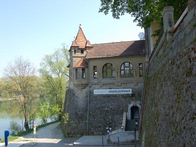 A stone building with a red-tiled turret and arched windows, featuring a sign reading "SCHNATL STADTGASTHAUS," situated next to a river with a path and a bridge in the background.