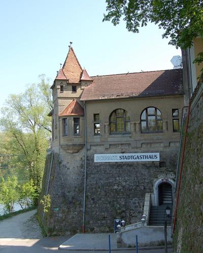 A stone building with a red-tiled turret and arched windows, featuring a sign reading "SCHNATL STADTGASTHAUS," situated next to a river with a path and a bridge in the background.