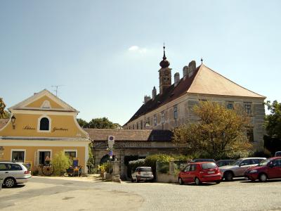 A yellow building with the text "Schloss Gasthaus" on its facade, situated next to a larger stone castle-like structure with a tower, with several cars parked in the foreground.