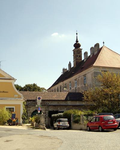 A yellow building with the text "Schloss Gasthaus" on its facade, situated next to a larger stone castle-like structure with a tower, with several cars parked in the foreground.