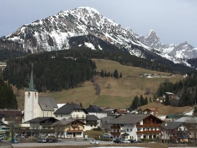 A view of the village of Filzmoos in the Salzburg region of Austria, featuring the white parish church with a green spire in the foreground and snow-capped mountains in the background.