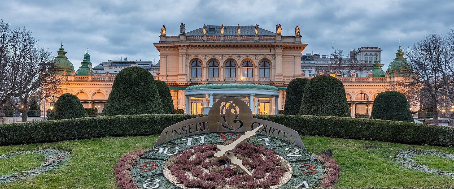 A large floral clock in a manicured garden with the text "UNSERE GARTEN" on its face, set against a grand neoclassical building with statues and green domes under a cloudy sky.