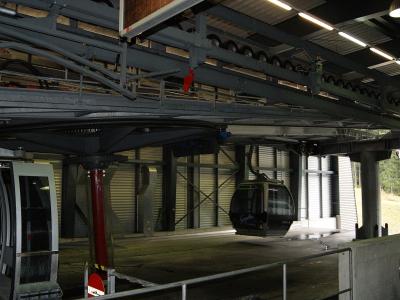 The image shows the interior of a cable car station, specifically the lower terminal of a gondola lift, featuring the mechanical track system, a red support pillar, and a cabin on the line.