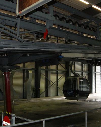 The image shows the interior of a cable car station, specifically the lower terminal of a gondola lift, featuring the mechanical track system, a red support pillar, and a cabin on the line.