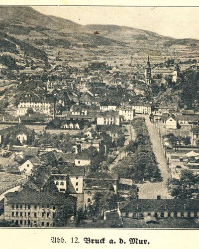 A vintage black-and-white panoramic photograph of a town nestled in a valley with hills in the background, featuring a prominent church spire and a long tree-lined avenue, with the caption "Abb. 12. Bruck a. d. Mur." at the bottom.