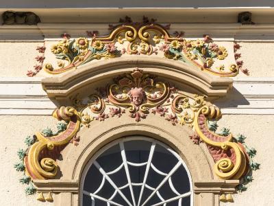 A close-up architectural detail of an ornate, Baroque-style window pediment featuring colorful stucco decorations, including floral motifs, scrolls, and a central cherub face, set against a cream-colored wall.