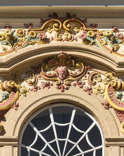 A close-up architectural detail of an ornate, Baroque-style window pediment featuring colorful stucco decorations, including floral motifs, scrolls, and a central cherub face, set against a cream-colored wall.